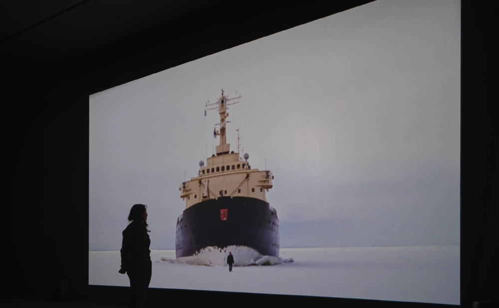 a woman watches a video projection of a boat on ice
